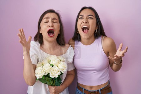 Foto de Madre e hija hispanas sosteniendo ramo de flores blancas locas y locas gritando y gritando con expresión agresiva y brazos levantados. concepto de frustración. - Imagen libre de derechos