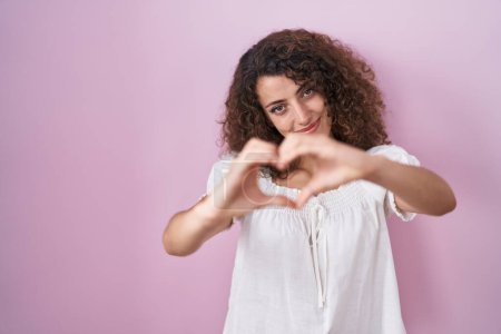 Foto de Mujer hispana con el pelo rizado de pie sobre fondo rosa sonriendo en el amor haciendo forma de símbolo del corazón con las manos. concepto romántico. - Imagen libre de derechos