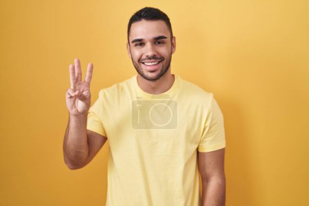 Téléchargez les photos : Young hispanic man standing over yellow background showing and pointing up with fingers number three while smiling confident and happy. - en image libre de droit