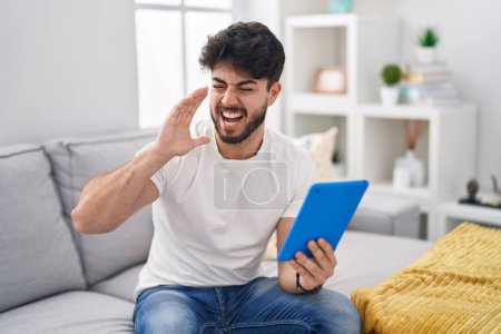 Foto de Hombre hispano con barba usando touchpad sentado en el sofá gritando y gritando fuerte a un lado con la mano en la boca. concepto de comunicación. - Imagen libre de derechos