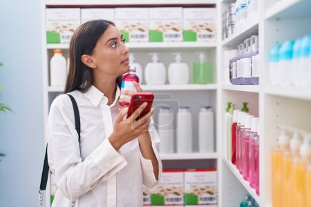 Téléchargez les photos : Jeune belle hispanique femme client en utilisant smartphone tenant bouteille de médicament à la pharmacie - en image libre de droit