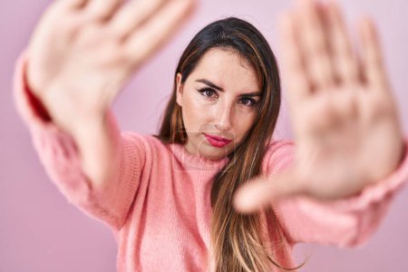 Foto de Mujer hispana joven de pie sobre fondo rosa haciendo marco usando las manos palmas y los dedos, perspectiva de la cámara - Imagen libre de derechos