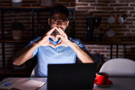 Photo for Hispanic man with beard using laptop at night smiling in love doing heart symbol shape with hands. romantic concept. - Royalty Free Image