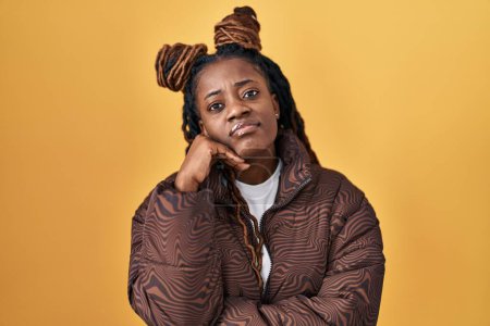 Photo for African woman with braided hair standing over yellow background with hand on chin thinking about question, pensive expression. smiling with thoughtful face. doubt concept. - Royalty Free Image