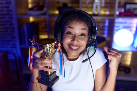 Téléchargez les photos : Jeune femme arabe jouant à des jeux vidéo tenant trophée criant fier, célébrant la victoire et le succès très excité avec le bras levé - en image libre de droit