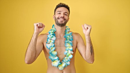 Photo for Young hispanic man tourist wearing hawaiian lei standing shirtless with winner gesture over isolated yellow background - Royalty Free Image