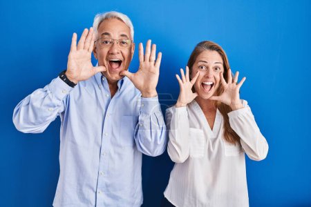Téléchargez les photos : Couple hispanique du Moyen Age debout sur fond bleu souriant gai jouer jeter un coup d'oeil un boo avec les mains montrant le visage. surpris et sortis - en image libre de droit