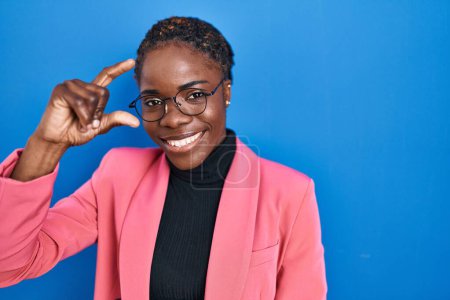 Photo for Beautiful black woman standing over blue background smiling and confident gesturing with hand doing small size sign with fingers looking and the camera. measure concept. - Royalty Free Image