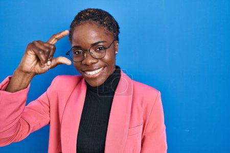 Photo for Beautiful black woman standing over blue background smiling and confident gesturing with hand doing small size sign with fingers looking and the camera. measure concept. - Royalty Free Image