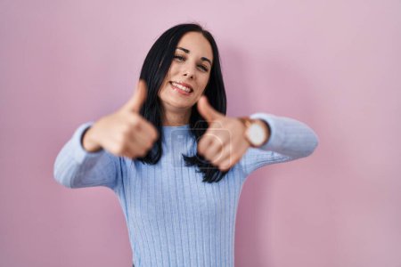 Foto de Mujer hispana de pie sobre fondo rosa aprobando hacer gesto positivo con la mano, pulgares hacia arriba sonriendo y feliz por el éxito. gesto ganador. - Imagen libre de derechos