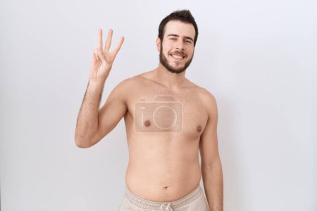 Foto de Joven hombre hispano de pie sin camisa sobre fondo blanco mostrando y señalando con los dedos número tres mientras sonríe confiado y feliz. - Imagen libre de derechos