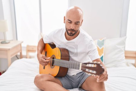 Photo for Young bald man playing classical guitar sitting on bed at bedroom - Royalty Free Image