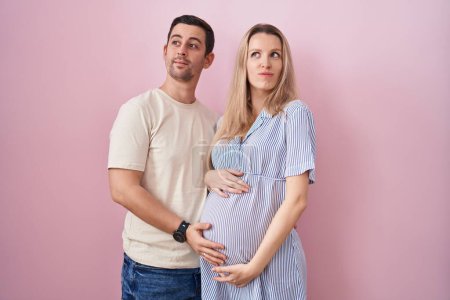 Photo for Young couple expecting a baby standing over pink background smiling looking to the side and staring away thinking. - Royalty Free Image