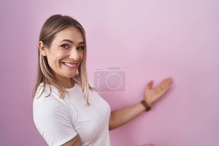 Foto de Mujer rubia caucásica de pie sobre fondo rosa invitando a entrar sonriendo natural con la mano abierta - Imagen libre de derechos