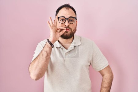 Foto de Hombre hispano de talla grande con barba de pie sobre fondo rosado boca y labios cerrados como cremallera con los dedos. secreto y silencioso, tabú hablando - Imagen libre de derechos