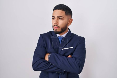 Téléchargez les photos : Jeune homme hispanique portant costume d'affaires et cravate regardant sur le côté avec les bras croisés convaincu et confiant - en image libre de droit