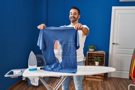 Photo for Young hispanic man with beard ironing holding burned iron shirt at laundry room smiling and laughing hard out loud because funny crazy joke. - Royalty Free Image