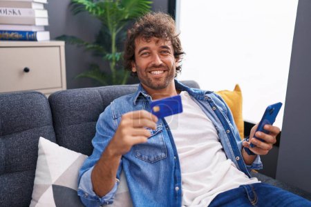 Foto de Joven hombre hispano usando teléfono inteligente y tarjeta de crédito sentado en el sofá en casa - Imagen libre de derechos