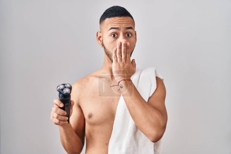 Photo for Young hispanic man holding electric razor covering mouth with hand, shocked and afraid for mistake. surprised expression - Royalty Free Image