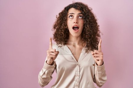 Téléchargez les photos : Hispanic woman with curly hair standing over pink background amazed and surprised looking up and pointing with fingers and raised arms. - en image libre de droit