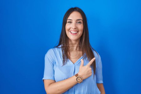 Photo for Young brunette woman standing over blue background cheerful with a smile on face pointing with hand and finger up to the side with happy and natural expression - Royalty Free Image