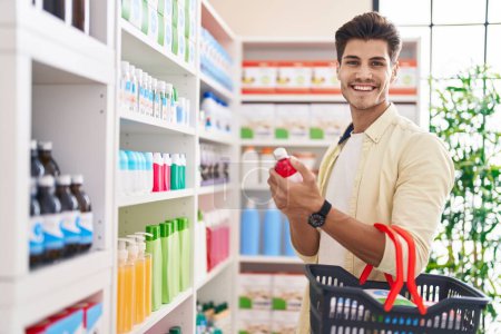 Téléchargez les photos : Jeune homme hispanique client souriant confiant tenant bouteille de médicaments et panier de marché à la pharmacie - en image libre de droit
