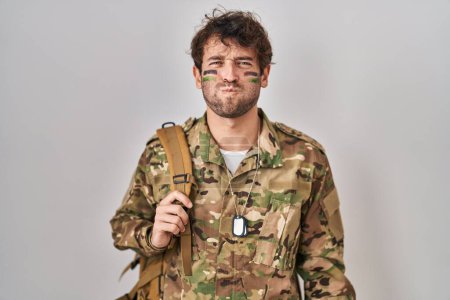 Photo for Hispanic young man wearing camouflage army uniform puffing cheeks with funny face. mouth inflated with air, crazy expression. - Royalty Free Image