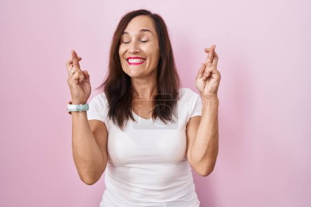 Téléchargez les photos : Femme brune du moyen âge debout sur fond rose doigt gestuel croisé souriant avec espoir et les yeux fermés. chance et concept superstitieux. - en image libre de droit