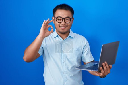 Téléchargez les photos : Chinois jeune homme utilisant ordinateur portable souriant positif faire ok signe avec la main et les doigts. expression réussie. - en image libre de droit