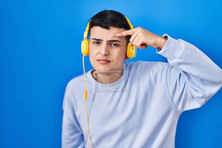Photo for Non binary person listening to music using headphones pointing unhappy to pimple on forehead, ugly infection of blackhead. acne and skin problem - Royalty Free Image