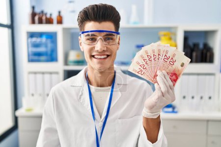 Foto de Joven hombre hispano trabajando en laboratorio científico sosteniendo shekels buscando positivo y feliz de pie y sonriendo con una sonrisa confiada mostrando los dientes - Imagen libre de derechos