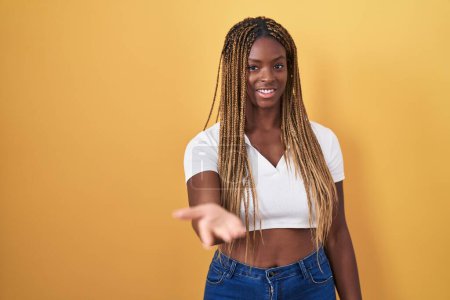 Téléchargez les photos : Femme afro-américaine avec des cheveux tressés debout sur fond jaune souriant joyeuse offrant main de paume donnant assistance et acceptation. - en image libre de droit