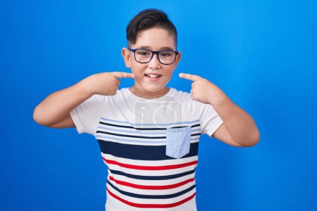 Téléchargez les photos : Jeune enfant hispanique debout sur fond bleu souriant gai montrant et pointant avec les doigts dents et la bouche. concept de santé dentaire. - en image libre de droit