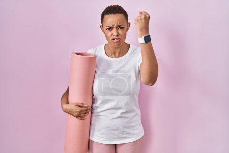 Téléchargez les photos : Belle femme afro-américaine tenant tapis de yoga en colère et fou levant le poing frustré et furieux tout en criant avec colère. rage et concept agressif. - en image libre de droit