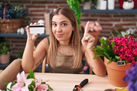 Téléchargez les photos : Jeune femme blonde travaillant dans un magasin de fleuristes tenant une carte de crédit souriant avec une idée ou une question pointant du doigt avec un visage heureux, numéro un - en image libre de droit