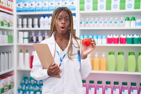 Téléchargez les photos : Femme afro-américaine aux cheveux tressés travaillant à la pharmacie pharmacie effrayée et choquée par l'expression surprise et étonnée, la peur et le visage excité. - en image libre de droit
