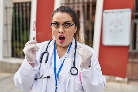 Foto de Joven doctora hispana haciendo prueba nasal de infección por coronavirus asustada y sorprendida con expresión sorpresiva y asombrada, miedo y cara excitada. - Imagen libre de derechos