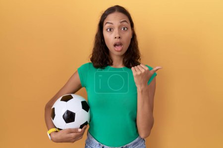 Foto de Mujer hispana joven sosteniendo la pelota sorprendida señalando con el dedo de la mano hacia un lado, boca abierta expresión asombrada. - Imagen libre de derechos