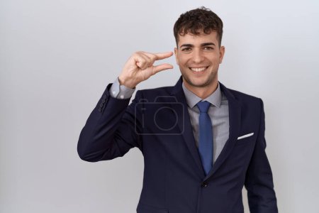 Téléchargez les photos : Jeune homme d'affaires hispanique portant costume et cravate geste souriant et confiant avec la main faisant signe de petite taille avec les doigts à la recherche et la caméra. concept de mesure. - en image libre de droit
