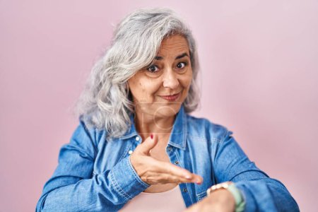 Foto de Mujer de mediana edad con el pelo gris de pie sobre el fondo rosa en la prisa que apunta a ver el tiempo, la impaciencia, molesto y enojado por retraso plazo - Imagen libre de derechos
