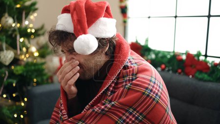 Photo for Young hispanic man feeling cold covered with blanket sitting on sofa by christmas tree at home - Royalty Free Image