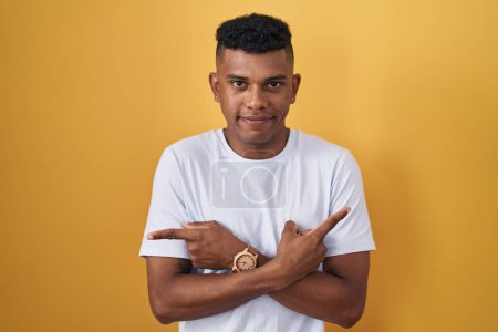 Téléchargez les photos : Young hispanic man standing over yellow background pointing to both sides with fingers, different direction disagree - en image libre de droit