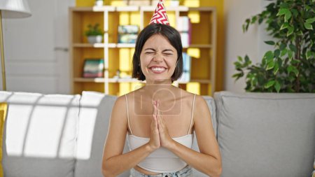 Téléchargez les photos : Jeune belle femme hispanique portant un chapeau d'anniversaire assis sur le canapé priant à la maison - en image libre de droit
