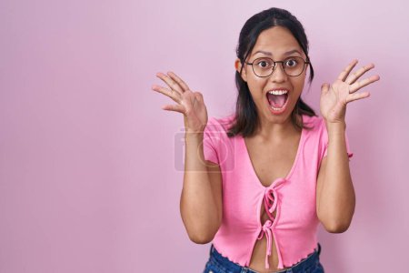 Photo for Hispanic young woman standing over pink background wearing glasses celebrating crazy and amazed for success with arms raised and open eyes screaming excited. winner concept - Royalty Free Image