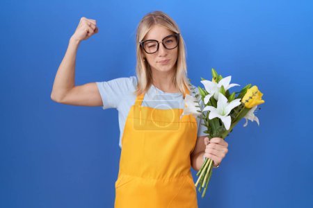 Foto de Mujer joven caucásica con delantal florista sosteniendo flores persona fuerte mostrando el músculo del brazo, confiado y orgulloso de poder - Imagen libre de derechos