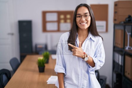 Photo for Young hispanic woman at the office cheerful with a smile on face pointing with hand and finger up to the side with happy and natural expression - Royalty Free Image