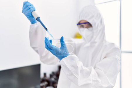 Téléchargez les photos : Jeune femme blonde scientifique portant l'uniforme de sécurité versant du liquide sur l'échantillon au laboratoire - en image libre de droit