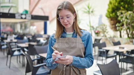 Foto de Mujer joven rubia camarera tomando notas en la terraza de la cafetería - Imagen libre de derechos