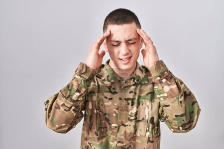 Foto de Joven vestido con camuflaje uniforme del ejército con la mano en la cabeza para el dolor en la cabeza porque el estrés. padecer migraña. - Imagen libre de derechos