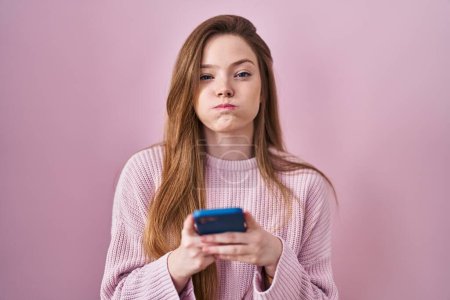 Téléchargez les photos : Jeune femme caucasienne à l'aide d'un smartphone tapant message joues gonflées avec drôle de visage. bouche gonflée d'air, attrapant l'air. - en image libre de droit